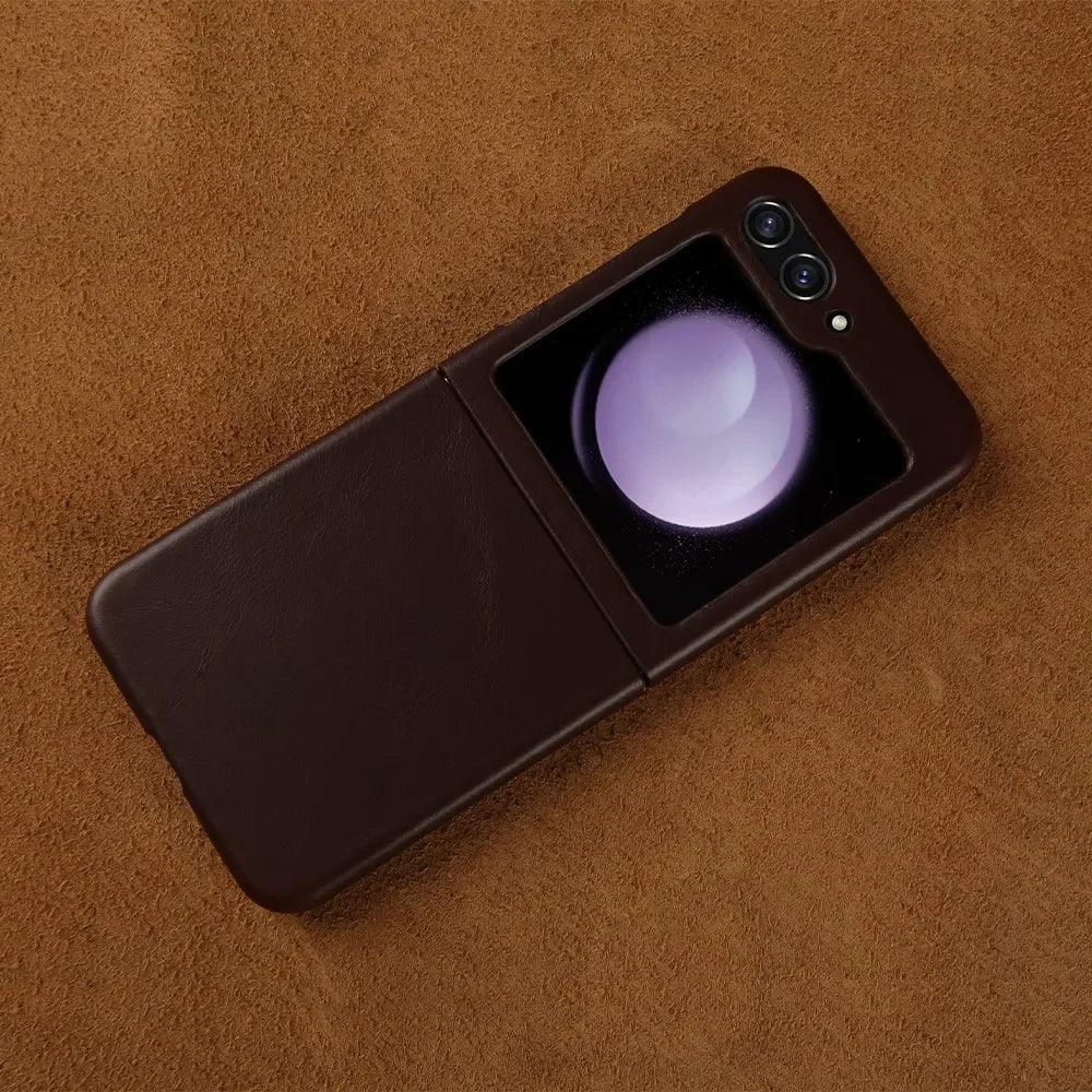 A brown Samsung Galaxy Z Flip foldable smartphone with dual rear cameras and a small external display showing a glowing orb, features genuine leather finish and is placed on a brown textured surface.