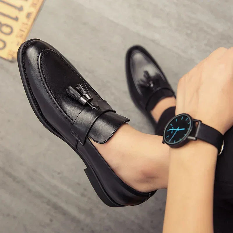 A person wearing black polished leather tassel loafers and a black wristwatch with a blue-accented face sits on a gray floor.