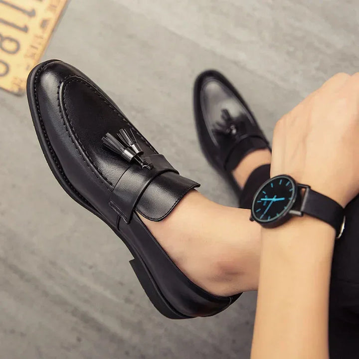 A person wearing black polished leather tassel loafers and a black wristwatch with a blue-accented face sits on a gray floor.