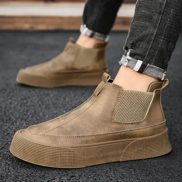 A person wearing brown slip-on suede sneakers with a cushioned footbed, thick textured soles, and elastic side panels, paired with dark, frayed hem jeans, stands on a black asphalt surface.
