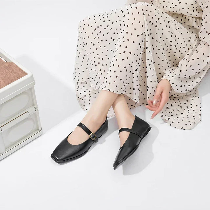 A person wearing a cream-colored, long, polka dot dress sits on the floor with legs extended, showcasing stylish black Mary Jane shoes with a buckle strap.