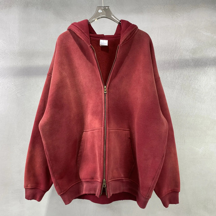 A spray-dyed vintage red zip-up hoodie with a relaxed fit and two front pockets hangs on a clear hanger against a light gray textured wall.