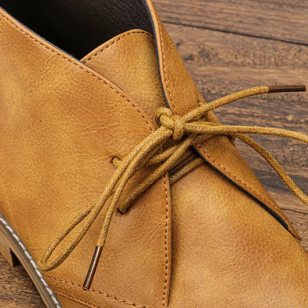 Close-up of a tan After-Ride Chukka leather shoe with untied laces resting on a wooden surface. The shoe showcases visible stitching details and metal aglets, highlighting its Western-inspired chukka design.