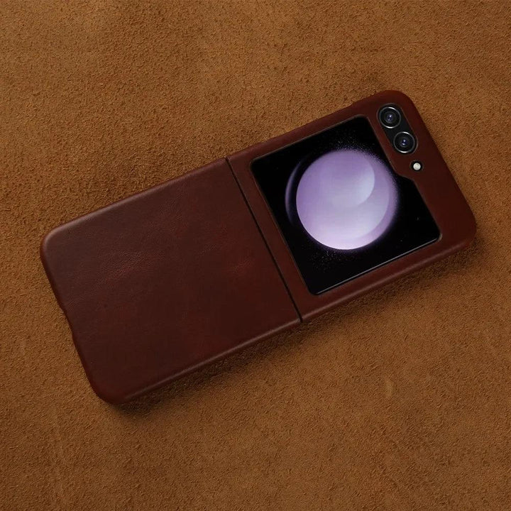 A Samsung Galaxy Z Flip in a genuine leather brown case rests on a suede surface. The phone is partially open, revealing its glowing external screen and dual camera lenses while staying protected with style.