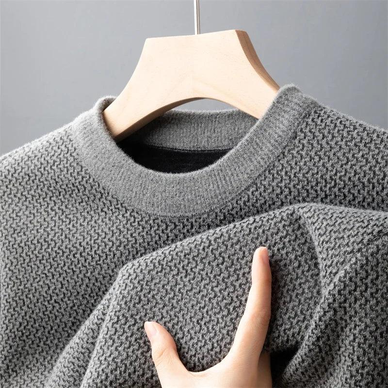 A hand is touching the sleeve of a relaxed fit sweater, crafted in textured, light gray knit, hanging on a wooden hanger against a plain gray background.
