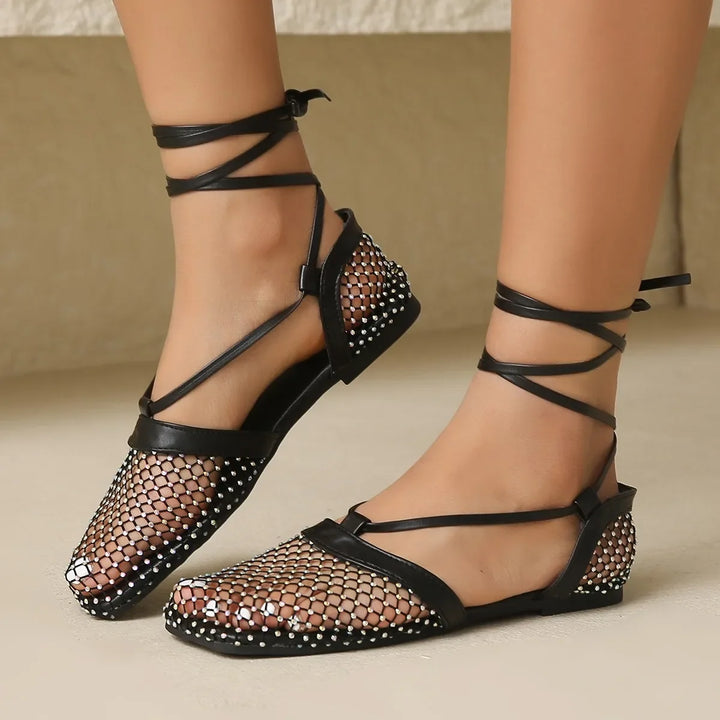A pair of feet wearing black flat shoes with a mesh upper and rhinestone detailing, featuring crisscross ankle straps tied around the ankles and a slip-resistant sole for added comfort.