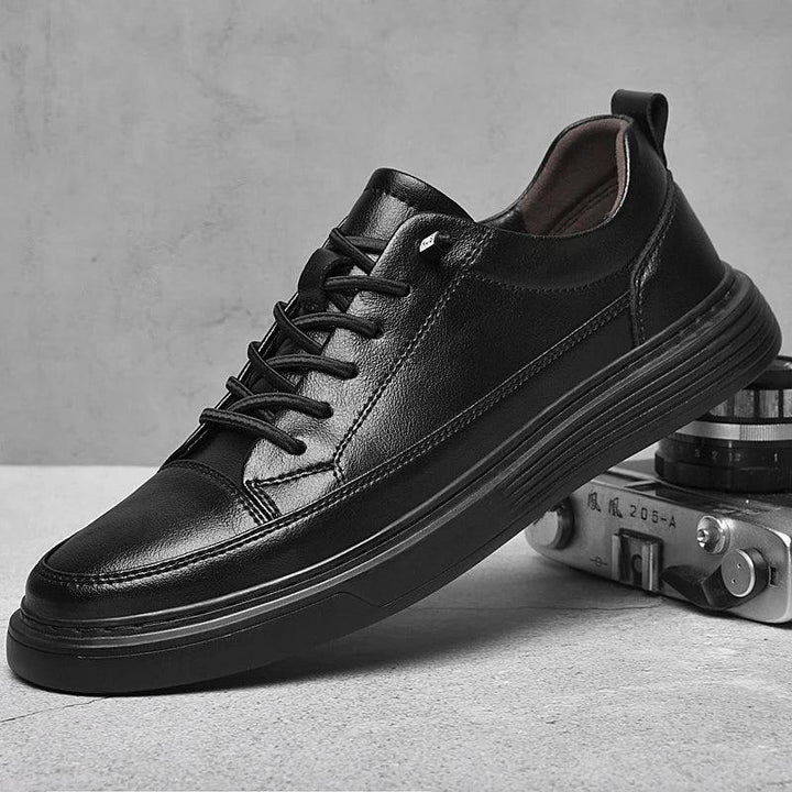A black genuine leather sneaker with black laces is propped against a vintage camera on a light surface, with a textured gray background. The shoe features a thick sole, minimal stitching details, and comfortable RunForm insoles.