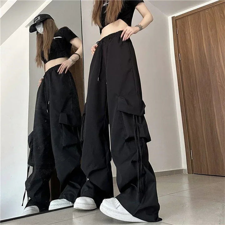 A person in a black crop top, high-waist black wide-leg cargo pants with cargo pockets, and white sneakers poses in front of a mirror. The outfit is finished with a black cap, standing on a light-colored floor near a wooden door.