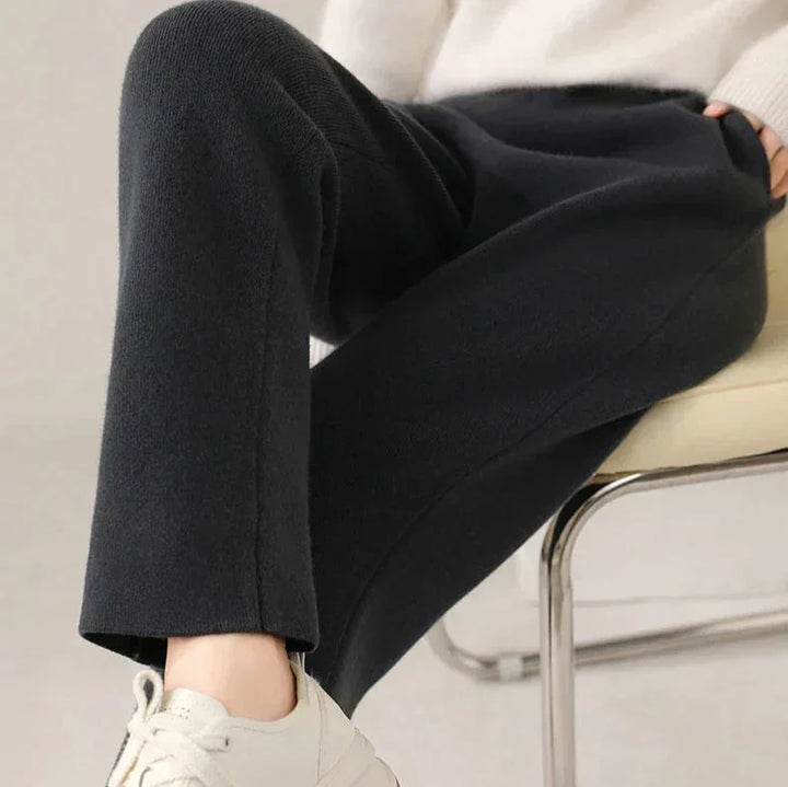 A person seated on a chair, wearing black pants with a slim tailored leg, a light-colored sweater, and white sneakers. The photo focuses on their legs and part of their hand resting on their lap.