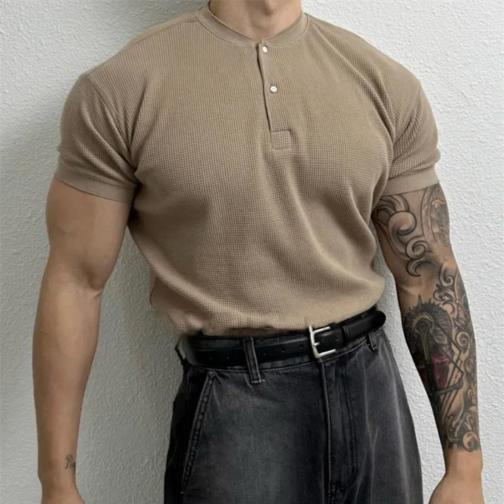 A person wearing a beige, short-sleeved, form-fitting shirt with a black belt and dark jeans stands against a white textured wall. Their right arm is tattooed, and their head is not visible in the image.
