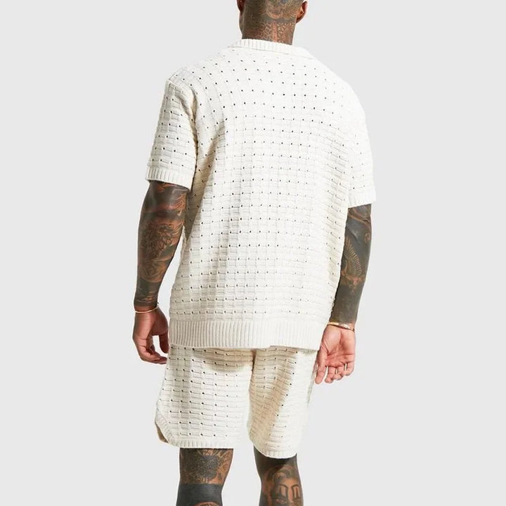 A man with tattooed arms and legs is shown from the back, wearing a breathable knit design off-white short-sleeve summer shirt and matching shorts. The background is light gray.