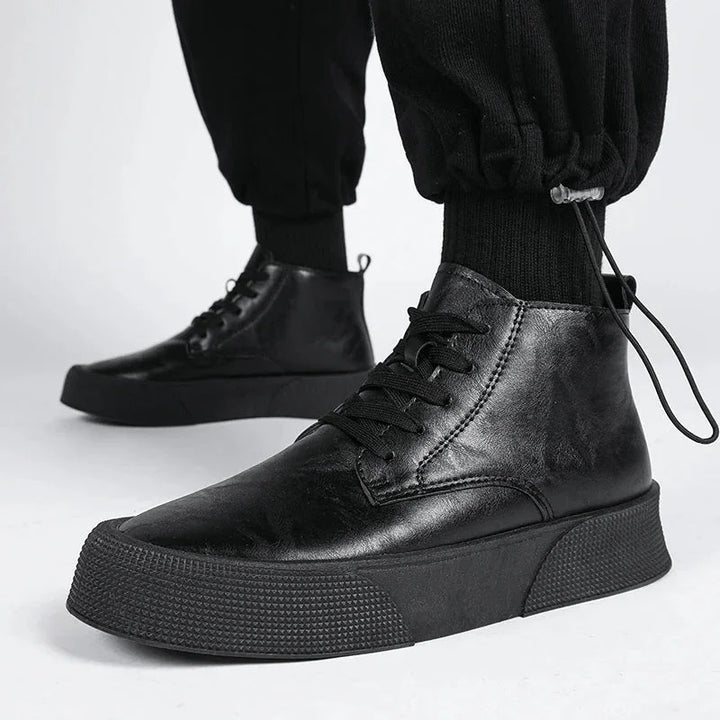 A person wears black vintage-inspired high-tops with thick soles and black laces, paired with black jogger pants. The photo focuses on the shoes cushioned footbed against a plain white background.