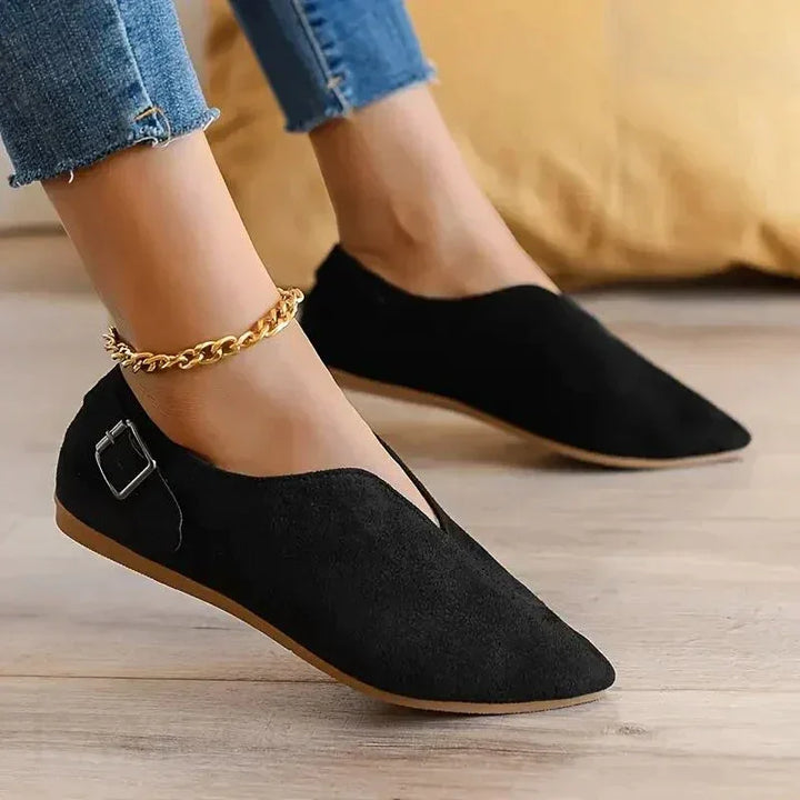 A person wearing black pointed toe shoes with a side buckle, slip-on design, and cushioned insole, paired with frayed hem blue jeans and a gold chain anklet, standing on a wooden floor with a yellow cushion in the background.