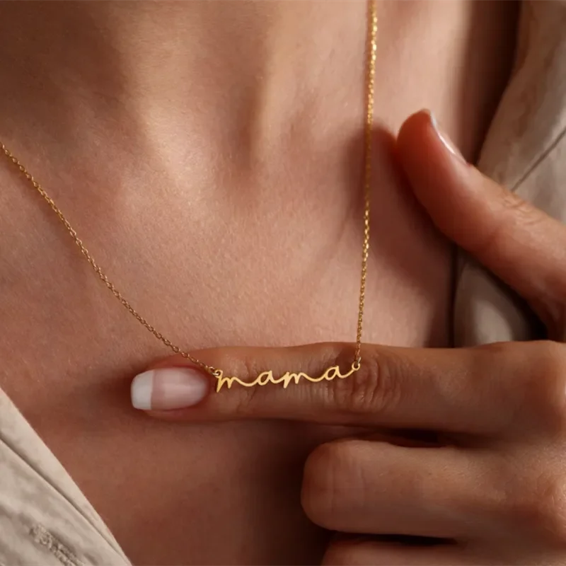 A person gently holds a gold-plated mama necklace with the word mama in cursive, resting on their chest. Their hand has neatly manicured nails and they are wearing a beige top—perfect as a Mothers Day necklace.