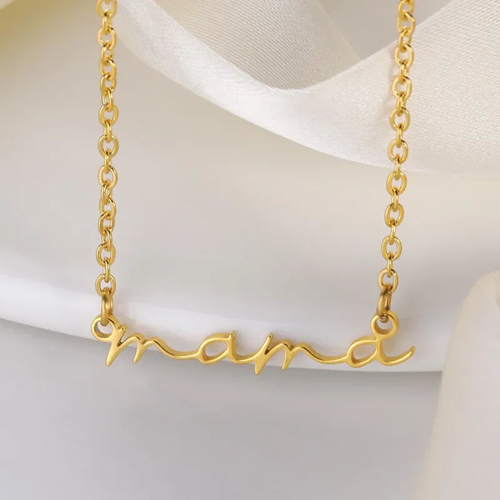 A gold-plated mama necklace with a delicate chain features a pendant that spells “mama” in cursive script, displayed against a soft, cream-colored fabric background.