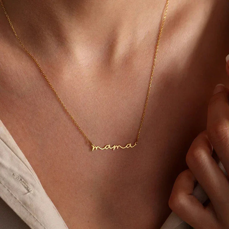 A close-up of a person wearing a delicate gold-plated mama necklace in cursive script. The Mothers Day necklace rests on their collarbone, paired with a light-colored top.