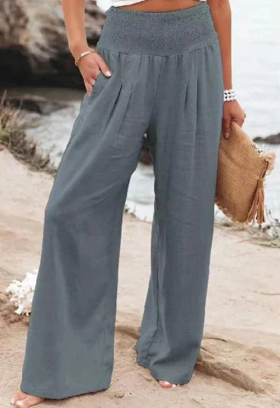 A person standing outdoors by the water, wearing loose-fitting, high waist summer pants and a white or light top, holding a woven clutch bag with tassels. Only the lower half of their body is visible.