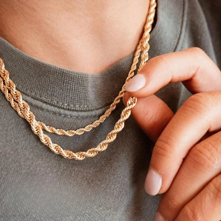 A person wearing a grey shirt holds one of two layered gold rope chain necklaces close to their neck with their fingers, showcasing the gold-toned jewelry. The focus is on the jewelry’s shine and adjustable sizing.