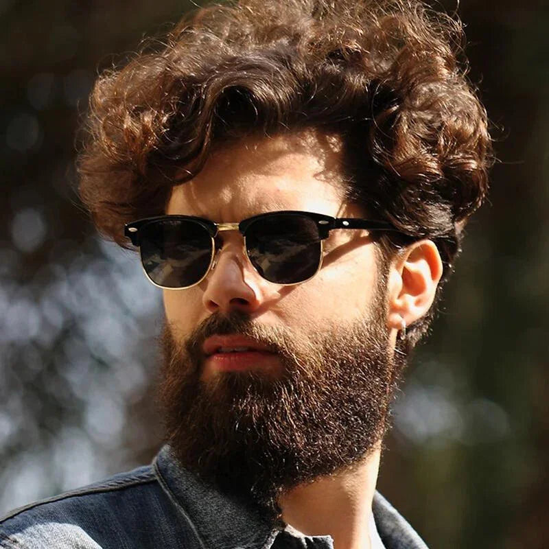 A man with curly brown hair, a full beard, and mustache wears vintage sunglasses with UV protection and a denim jacket outdoors. The background is blurred with hints of greenery and sunlight.