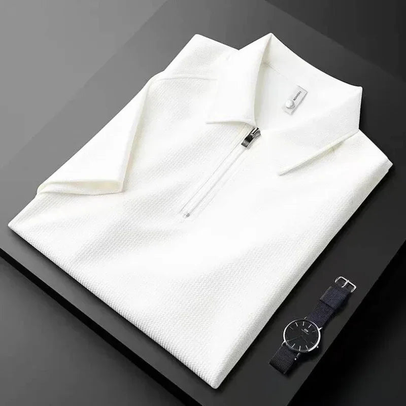 A neatly folded white mens silk polo shirt with a front zipper is placed on a black surface next to a minimalist black wristwatch with a black strap.