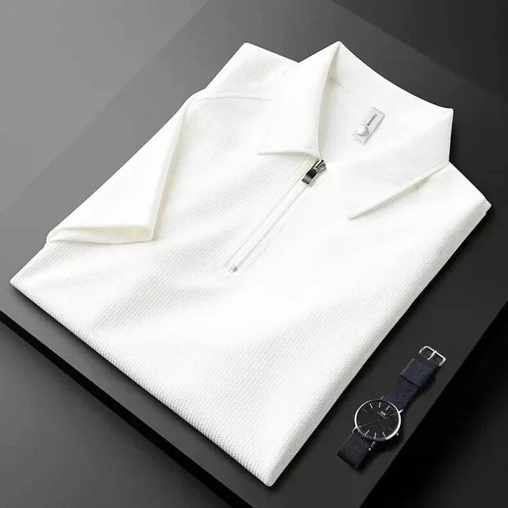A neatly folded white mens silk polo shirt with a front zipper is placed on a black surface next to a minimalist black wristwatch with a black strap.