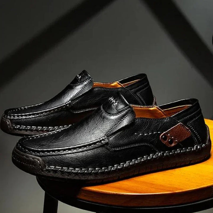 A pair of black leather loafers with detailed stitching and decorative side buckles, displayed on a round wooden stool under soft lighting.