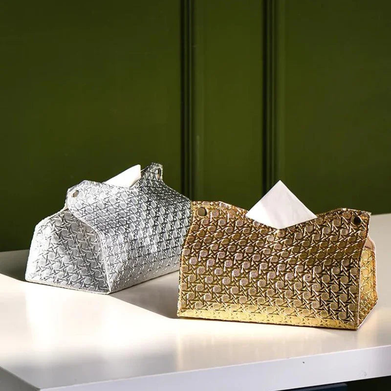 Two decorative tissue holders, the Glymmer Tissue Box in metallic silver and gold, sit on a white surface against a dark green background. Both feature a striking geometric pattern and hold tissues.