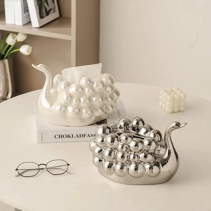 Two decorative snail-shaped containers with a bumpy, shiny surface sit on a table; one is a pearl white dispenser for tissues. Nearby are a pair of glasses, a small cube candle, a flower vase, and a book on the shelf in the background.