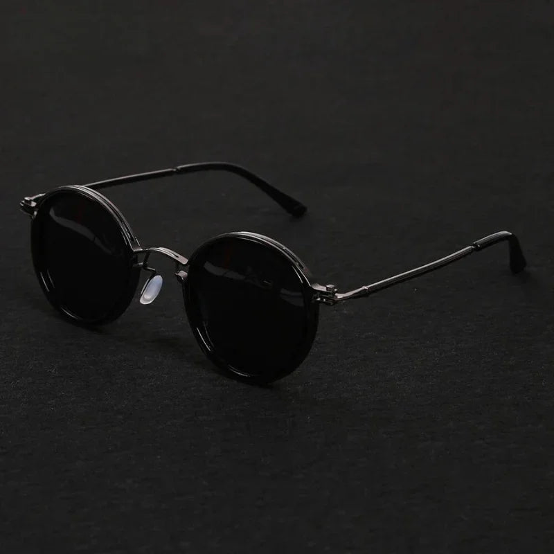 Round black Ruby Ray Sunglasses with dark lenses and thin metal frames are placed on a dark surface, their arms extended. These polarized sunglasses offer both style and enhanced UV400 protection for your eyes.