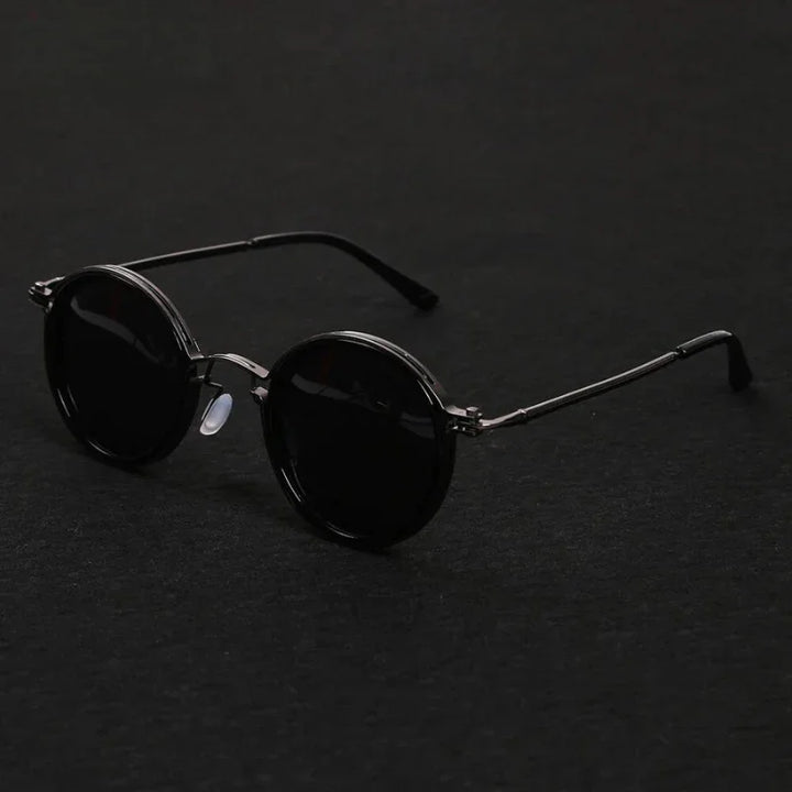 Round black Ruby Ray Sunglasses with dark lenses and thin metal frames are placed on a dark surface, their arms extended. These polarized sunglasses offer both style and enhanced UV400 protection for your eyes.