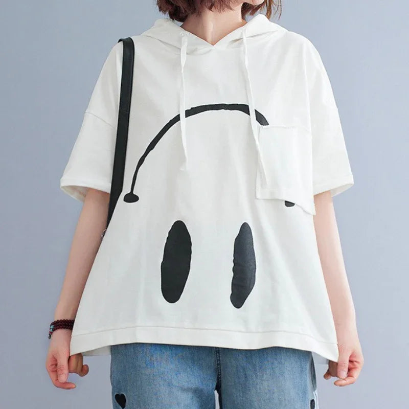 A person wears a loose white Monday Mood hoodie featuring a large black abstract face graphic and light blue jeans, standing against a plain background. The graphic shows two oval shapes and an arched line.