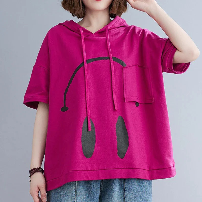 A person wearing a loose, bright pink Monday Mood short-sleeve hoodie with a large black abstract face design and a small chest pocket, paired with blue jeans. The spring colors pop, while the persons head is partially out of frame.