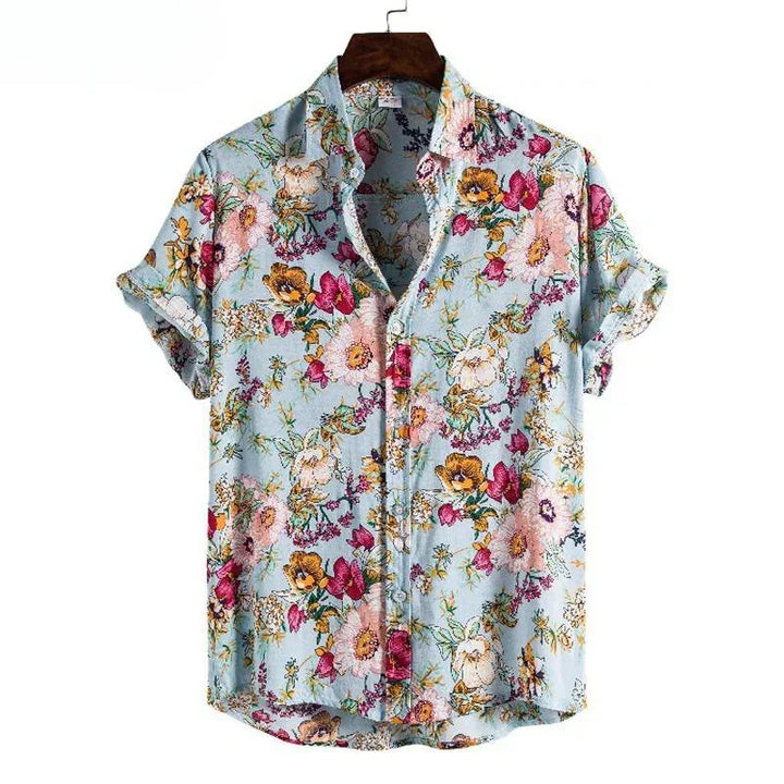 A short-sleeve, button-up, lightweight linen shirt with a light blue background features a vibrant floral pattern in pink, yellow, orange, and green. This vintage-inspired shirt is displayed on a hanger with the sleeves rolled up.