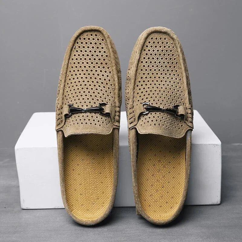A pair of light brown breathable loafers with metal bit accents, featuring innovative cushioning, displayed upright on white blocks against a gray background.