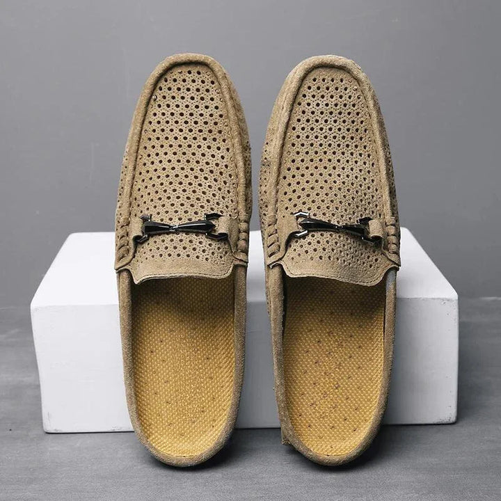 A pair of light brown breathable loafers with metal bit accents, featuring innovative cushioning, displayed upright on white blocks against a gray background.