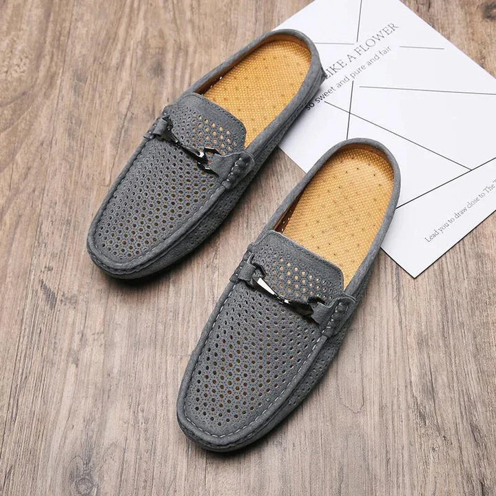 A pair of gray, perforated breathable loafers with tan insoles are placed on a wooden floor. One shoe rests partially on top of a white paper featuring light geometric lines and text.