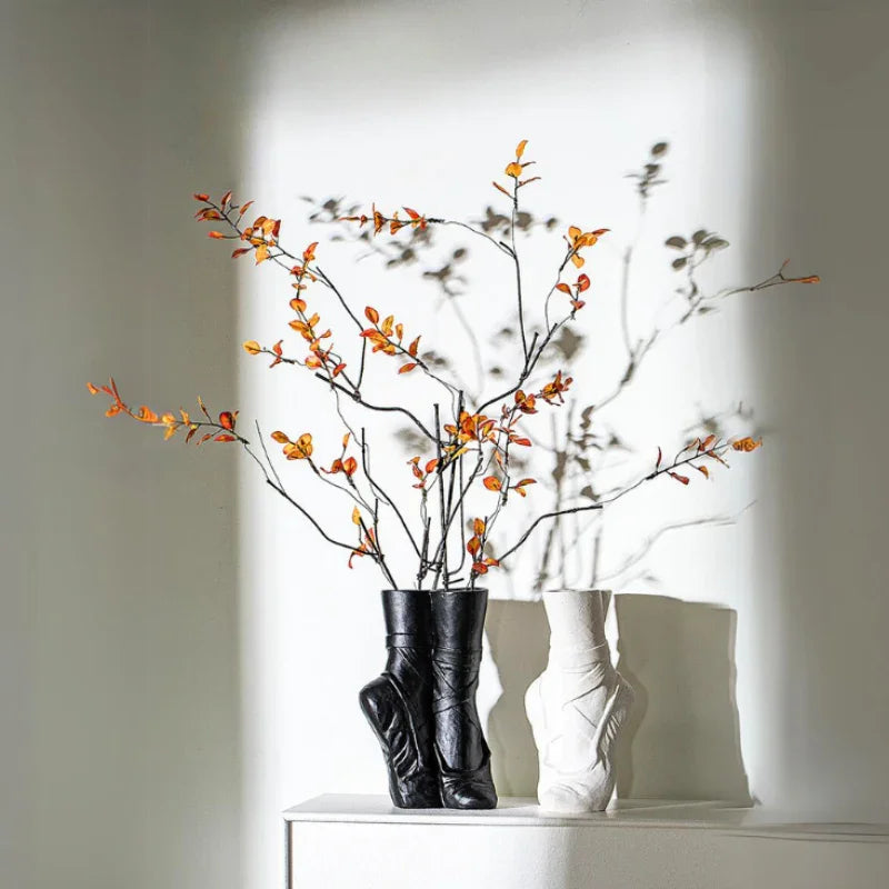 Two ballet-inspired ceramic vases, one black and one white, sit on a white surface. The black vase holds branches with small orange berries, casting dramatic shadows—perfect for contemporary chic décor.