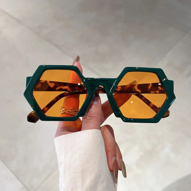 A hand with long, manicured nails holds trend-forward green hexagonal frame sunglasses with amber-tinted lenses and UV400 protection, against a light-colored tiled background.