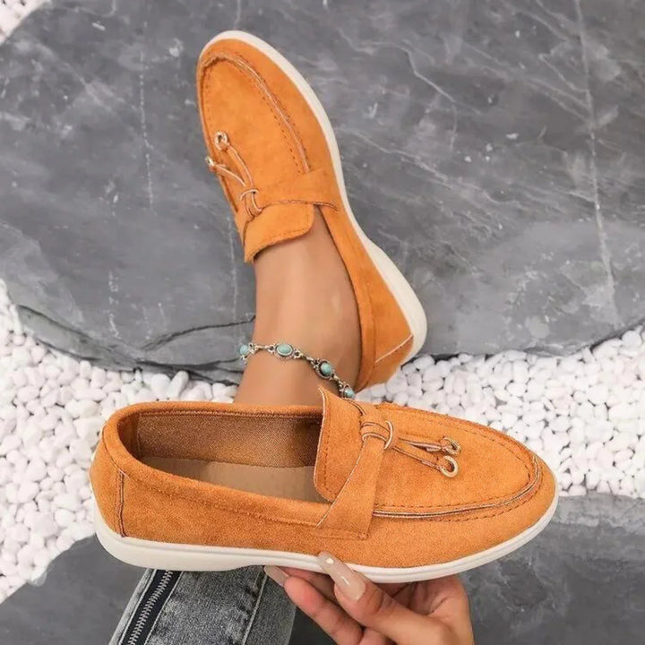 A person wearing chic suede moccasins with white soles sits on a stone surface with white pebbles, stylishly showcasing these streetwear shoes. They pair them with blue jeans and a beaded anklet for a trendy look.
