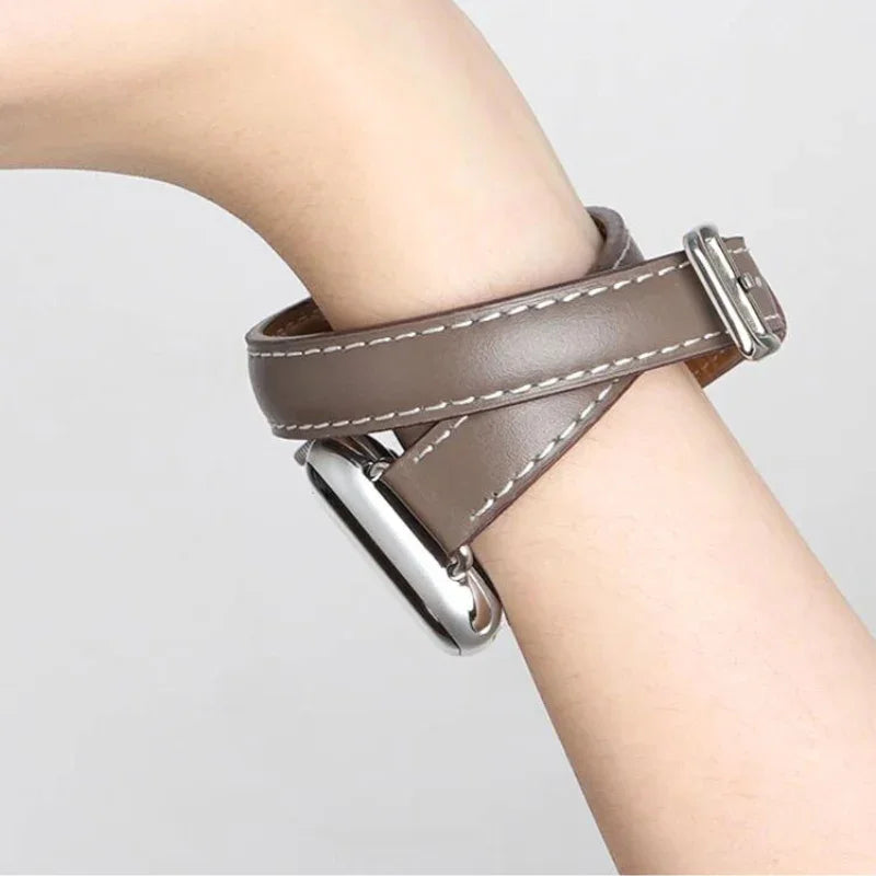 A close-up of a persons wrist wearing a brown genuine leather Apple Watch band with double wrap design, white stitching, and a silver rectangular watch face. The background is plain and light-colored.