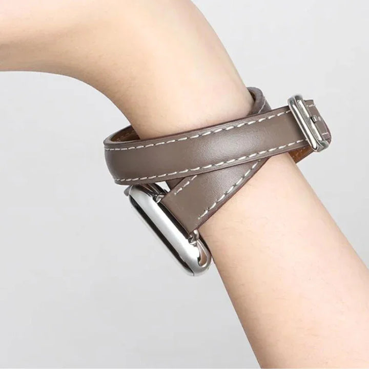 A close-up of a persons wrist wearing a brown genuine leather Apple Watch band with double wrap design, white stitching, and a silver rectangular watch face. The background is plain and light-colored.