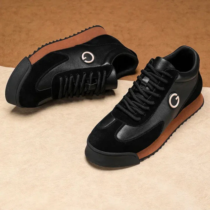 A pair of sleek sneakers in black with brown soles and a circular logo on the side, crafted from luxe leather suede, displayed on a beige and brown surface.