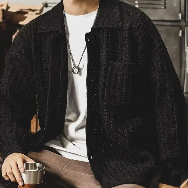 A person wearing a chunky knit black cardigan over a white shirt and brown pants sits holding a metal cup, with a silver necklace visible around their neck, capturing a relaxed heritage aesthetic.
