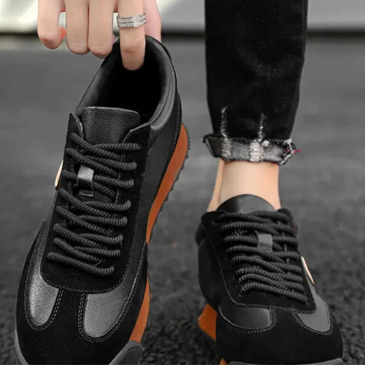 A person wearing sleek sneakers with thick black laces and brown soles stands on a dark surface, one hand holding the heel of the left shoe. The person’s jeans are slightly rolled at the ankles, adding an urban professional touch.
