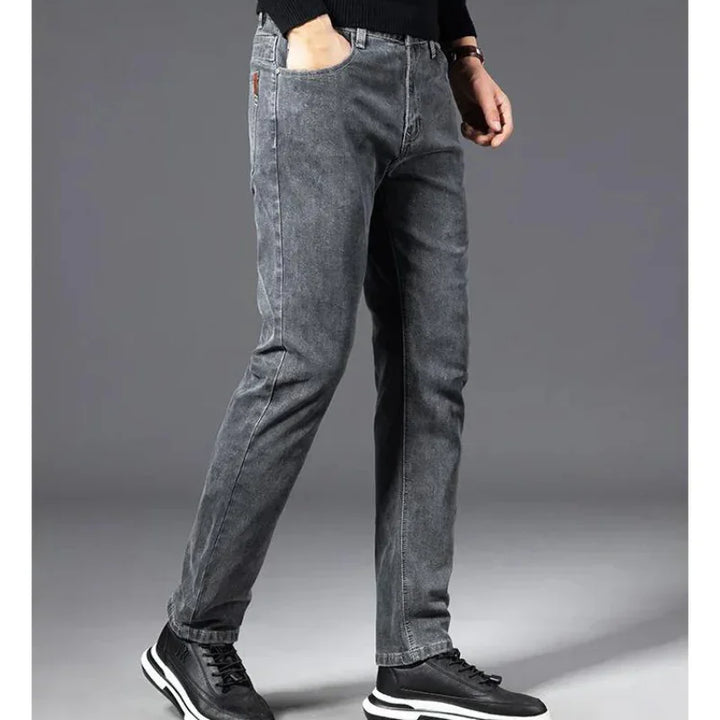 A person stands against a plain gray background, wearing heavyweight denim straight-leg jeans, a black long-sleeve shirt, and black sneakers with white soles. The persons head is not visible in the image.