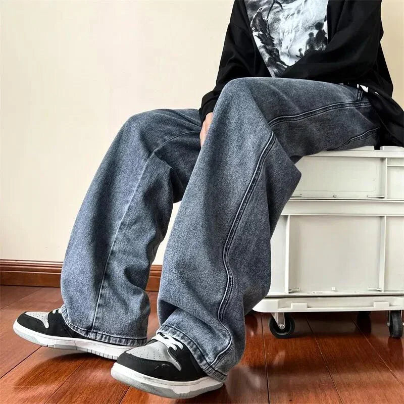 A person wearing relaxed fit, wide-legged blue jeans in a vintage wash, black and white sneakers, and a black long-sleeve shirt sits on a white storage box against a beige wall and wooden floor.
