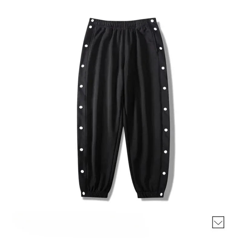 Black gym pants with an elastic waistband and cuffs, featuring white snap buttons along the outer sides of both legs. Made from moisture wicking fabric, the pants are laid flat on a white background.