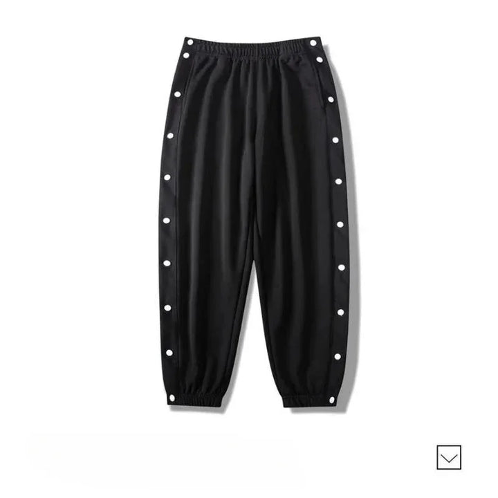 Black gym pants with an elastic waistband and cuffs, featuring white snap buttons along the outer sides of both legs. Made from moisture wicking fabric, the pants are laid flat on a white background.