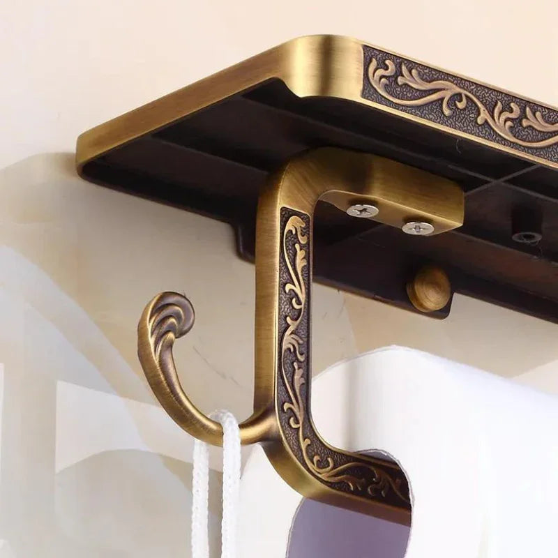 Close-up of an ornate wall hook with an antique brass finish and intricate detailing, mounted on a wall and holding a white cord. The hook, ideal for bathroom decor, features a shelf on top and vintage-style engravings.