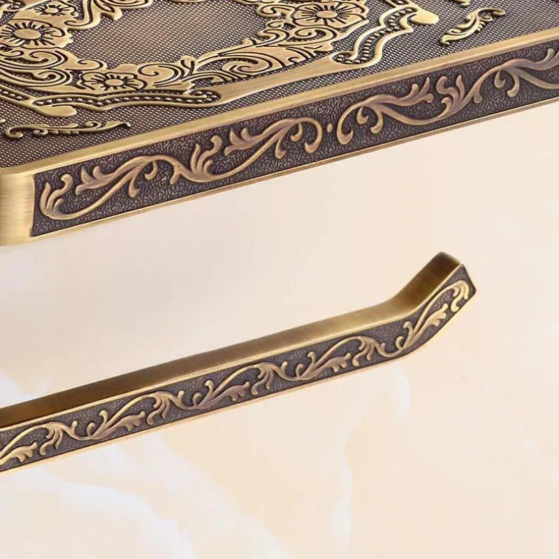A close-up of an ornate Victorian toilet paper holder with an antique brass finish, featuring intricate, embossed floral patterns and a matching decorative support bar against a light background—perfect for elegant bathroom decor.